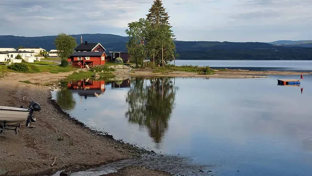 Rød hytte ved vannkanten med flytebrygge og speilbilde i vannet.