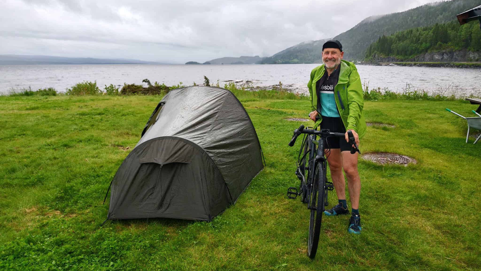Smilende syklist i regntøy ved telt nær vannkanten på gressplen.