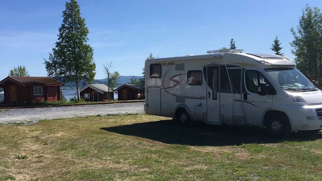 Bobil parkert på grønn gressplen ved campingplass med utsikt mot hytter, trær og vann i bakgrunnen.